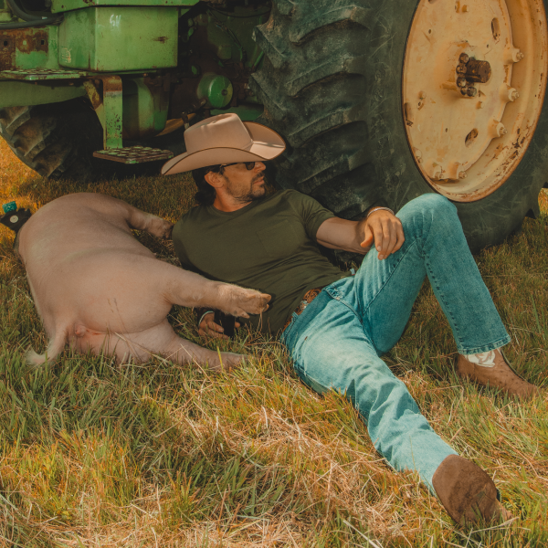 Propped against a tractor tire beside a napping pig, a man in an olive Shinesty tee, jeans and a cowboy hat