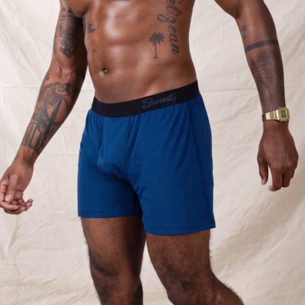 Tattooed man in royal blue Shinesty boxer shorts and a gold watch, front view against a canvas backdrop
