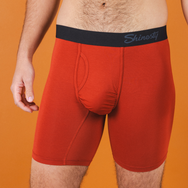 Rust-red Shinesty boxer briefs with a black logo waistband against an orange backdrop, legs reaching past mid-thigh