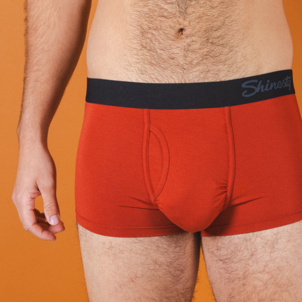 Short-legged rust-orange trunks against an orange wall, black Shinesty band at the waist and a front fly