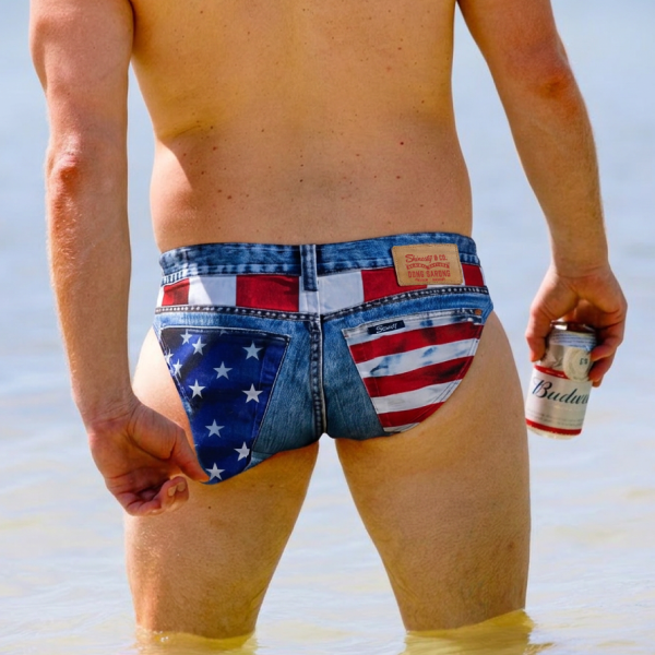 Man wading in shallow water in denim-print swim briefs with an American flag seat, beer can in hand