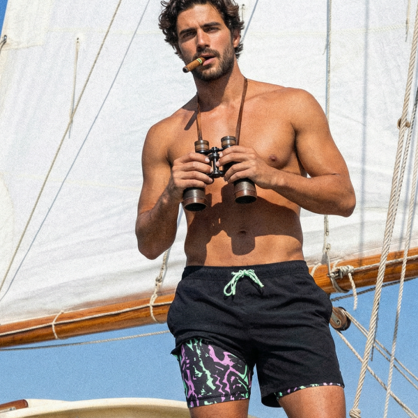 Man on a sailboat deck with binoculars and a cigar, black swim trunks with a neon-print liner at the hem
