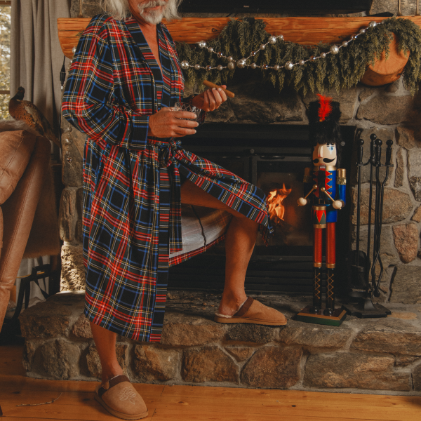 White-haired man in a red tartan robe seated by a stone fireplace with a nutcracker and garland