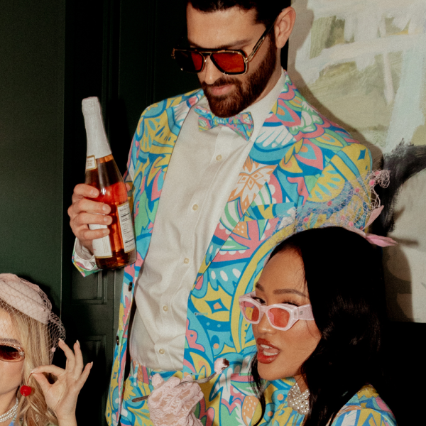 Man in red-tinted shades and a pastel pop-art party blazer holding a bottle of rosé, date in a matching dress beside him