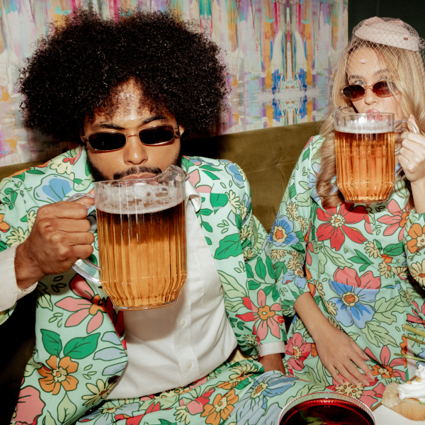 Friends in matching floral Shinesty party blazer and dress, each chugging beer from a full pitcher on a couch