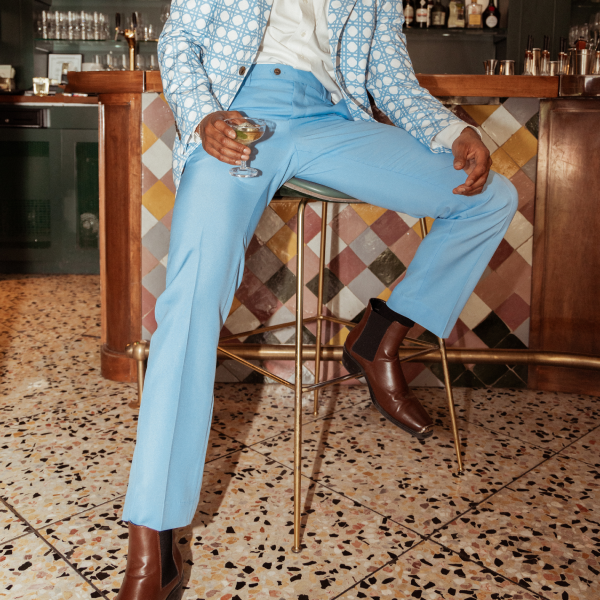 Man perched on a brass barstool in slim powder blue party suit trousers, patterned blazer and brown boots