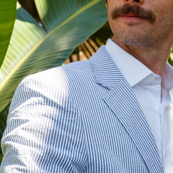Mustached man in a blue and white seersucker blazer over a white shirt, banana leaves behind