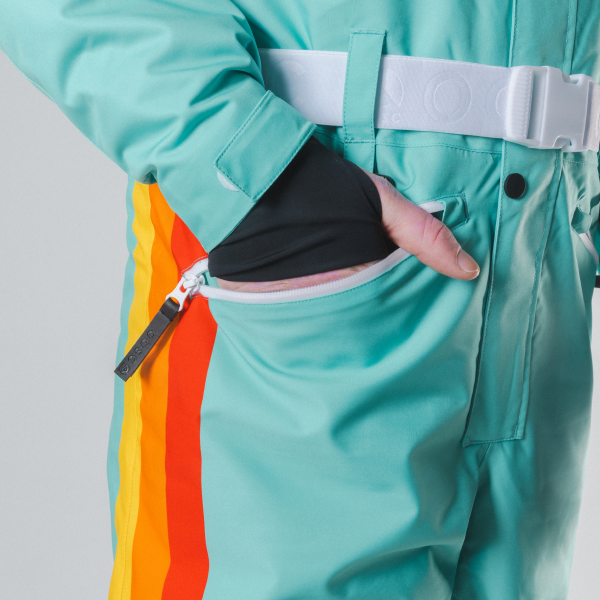 Hand in the zippered hip pocket of a teal ski suit, white belt buckled and rainbow stripe down the leg