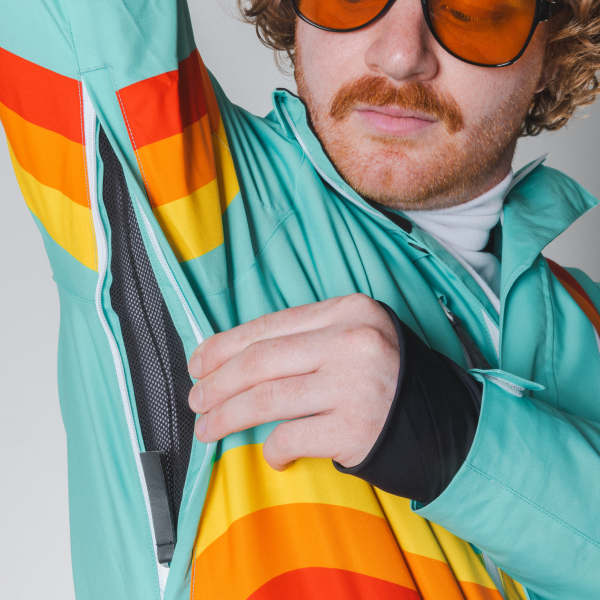 Man in orange sunglasses opening the mesh-lined underarm vent zipper on a teal Shinesty ski suit