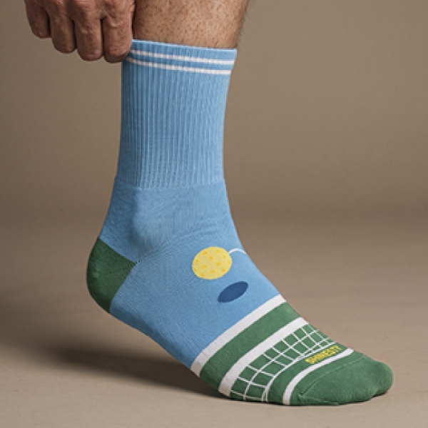 Sky blue quarter sock with a tennis court and ball design and a green toe, worn and lifted toward the camera