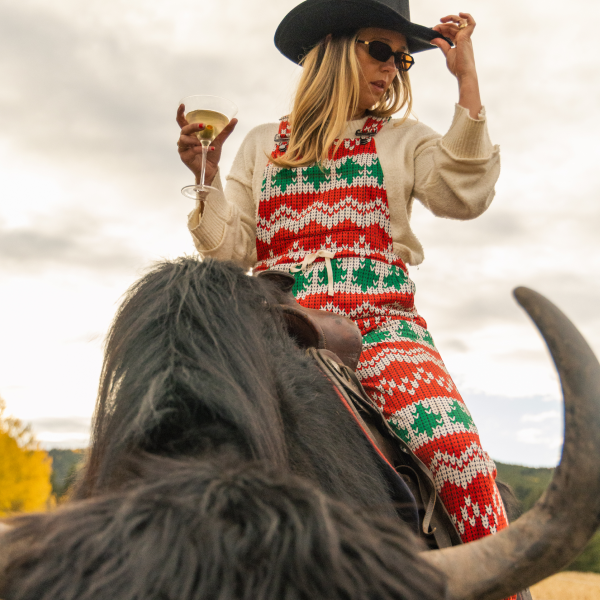 Woman in a black cowboy hat and holiday fair-isle pajamaralls raising a martini from the back of a yak
