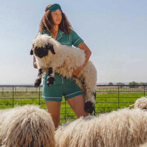 Green short-sleeve pajama set on a woman carrying a shaggy sheep across a pasture, sleep mask on her head