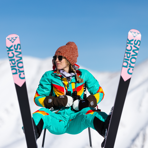 Skier in a teal rainbow-striped ski suit sitting in the snow between a pair of skis planted upright
