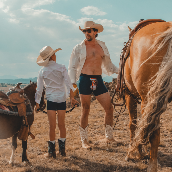 Father and son in white shirts, cowboy hats and boots wearing boxer briefs beside a palomino horse